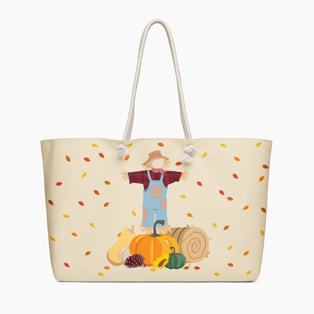 A cream-colored tote bag with white rope handles, featuring a printed autumn-themed illustration of a scarecrow standing behind a pumpkin with falling leaves. Staged on a light-grey background