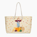 A cream-colored tote bag with white rope handles, featuring a printed autumn-themed illustration of a scarecrow standing behind a pumpkin with falling leaves. Staged on a light-grey background