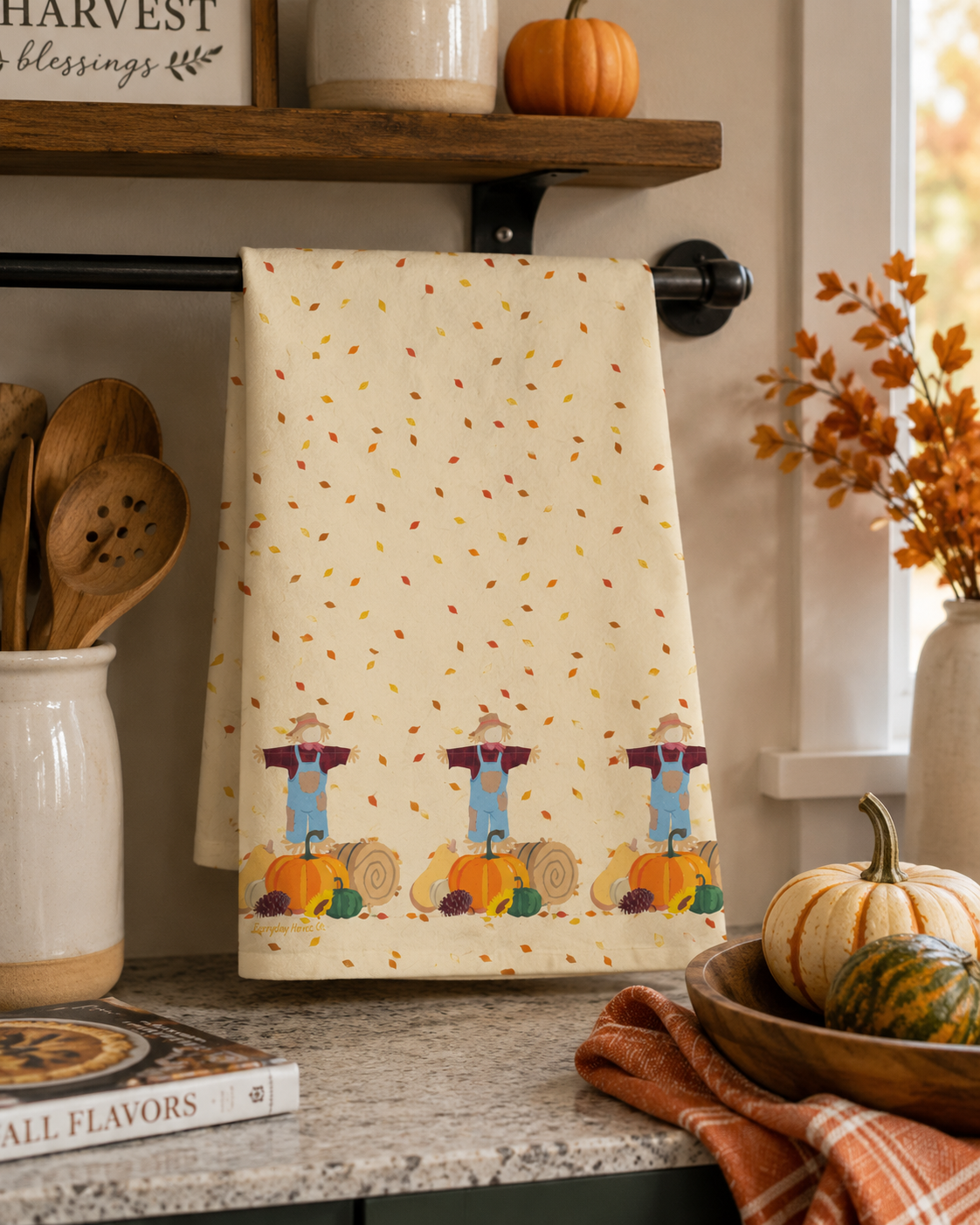 A kitchen towel with a fall harvest theme hangs on a black towel bar. The towel has a cream background with scattered orange, red, and yellow leaves, and three cartoon scarecrows standing behind pumpkins at the bottom.