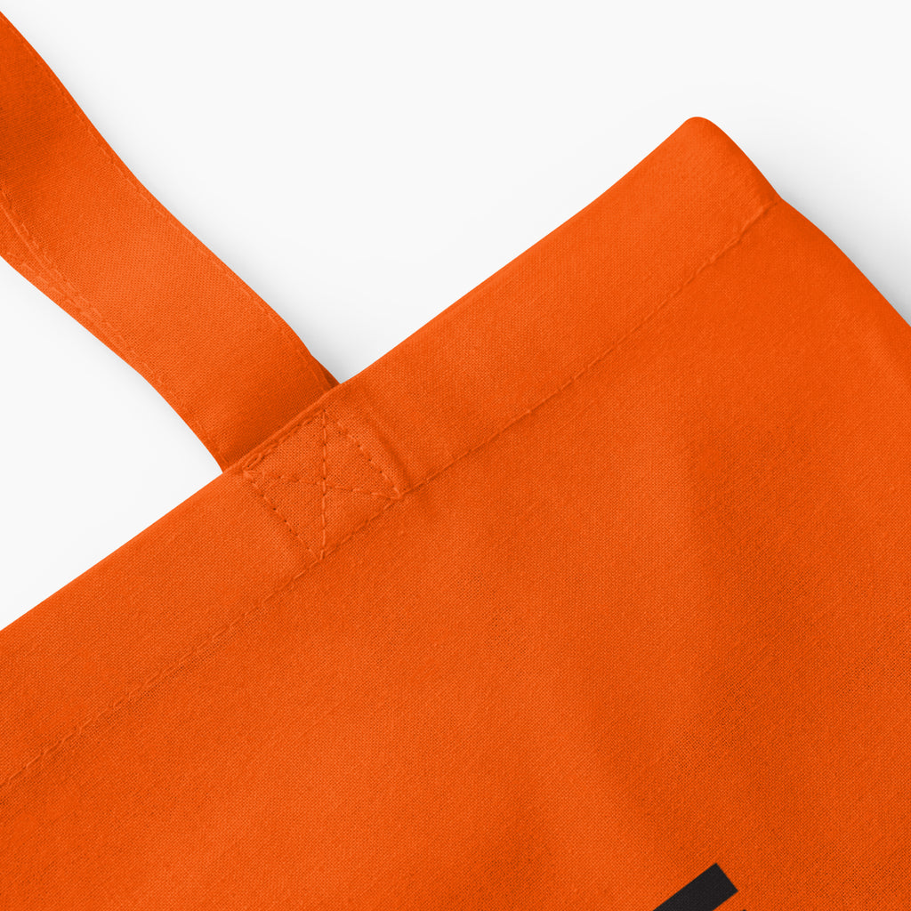 Close-up of an orange canvas tote bag showing the corner, stitched strap attachment on a light grey background.