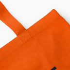 Close-up of an orange canvas tote bag showing the corner, stitched strap attachment on a light grey background.