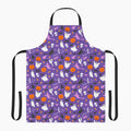 A purple apron with a black neck loop and waist ties, featuring an all-over Halloween pattern of orange pumpkins, white ghosts, black cats, and witches on a light gray background.
