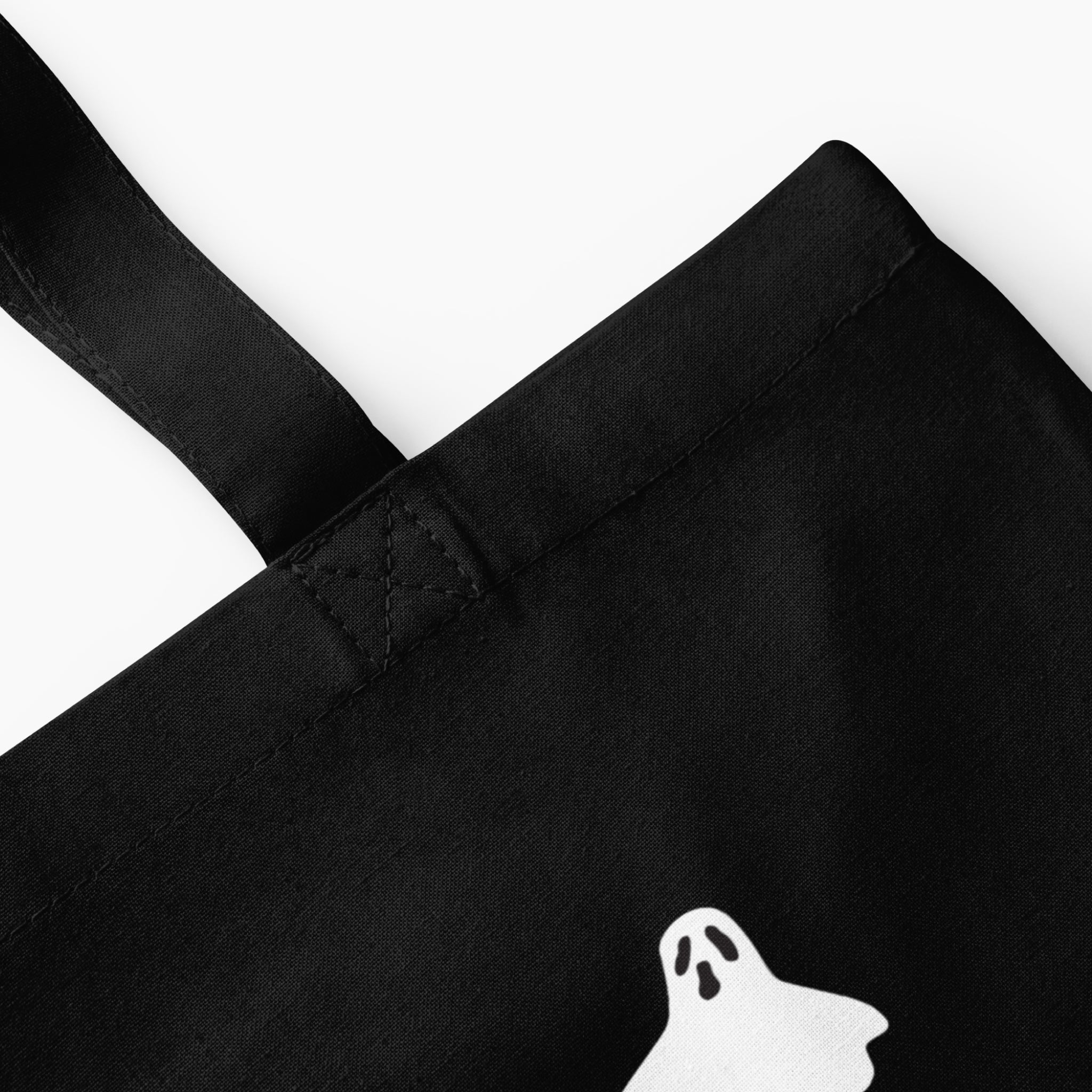 Close-up of a black fabric tote bag with a white ghost graphic and black straps on a light grey background.