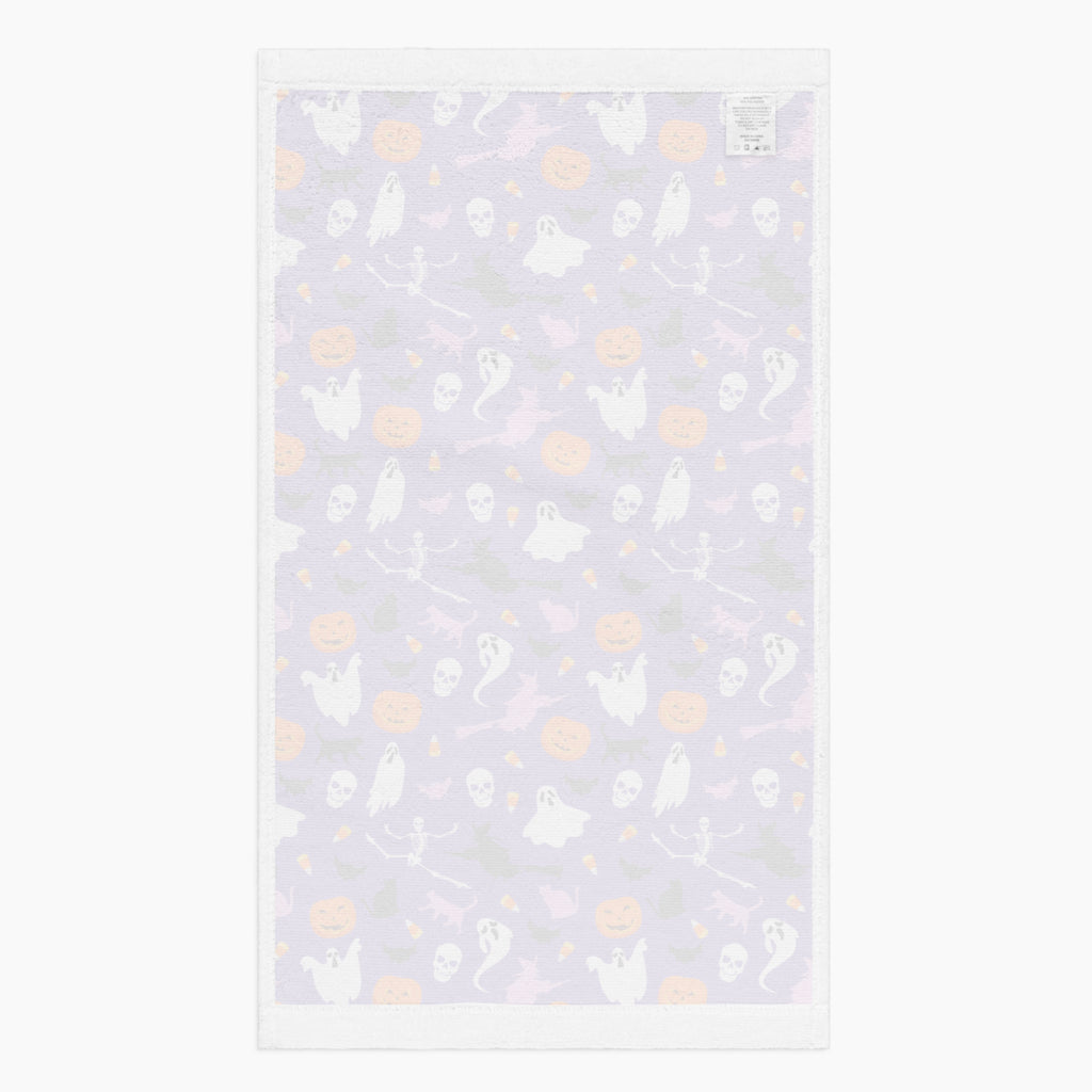 The white back of a rectangular cloth kitchen towel featuring a repeating Halloween pattern on a purple background, with white ghosts, black cats, orange pumpkins, and small candy corns.