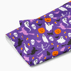 A folded cloth kitchen towel featuring a repeating Halloween pattern on a purple background, with white ghosts, black cats, orange pumpkins, and small candy corns.