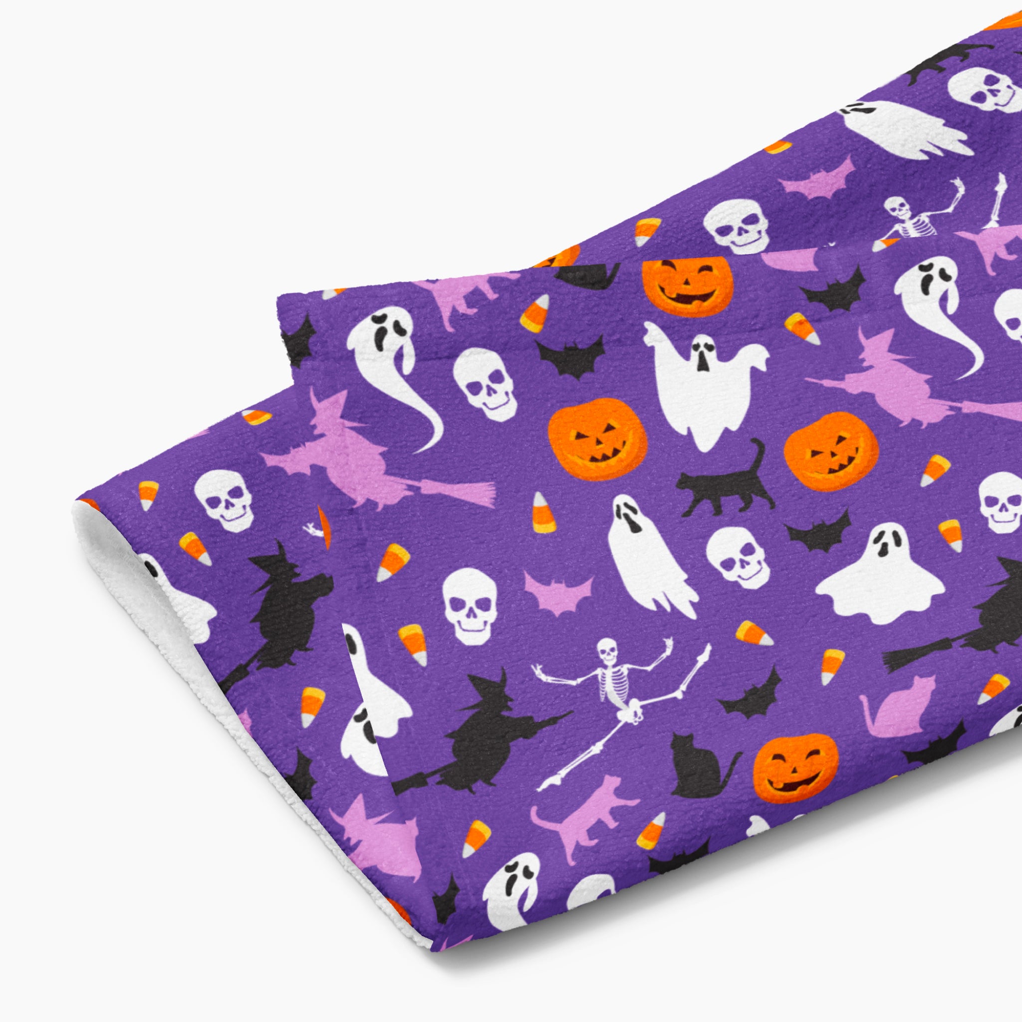 A folded cloth kitchen towel featuring a repeating Halloween pattern on a purple background, with white ghosts, black cats, orange pumpkins, and small candy corns.