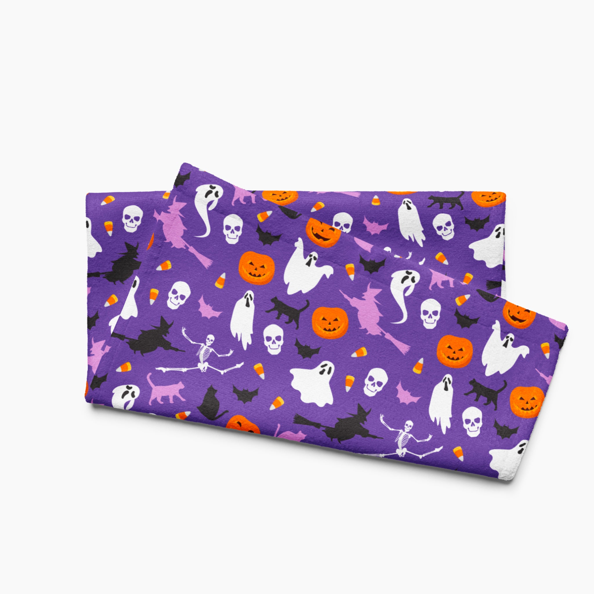 A folded cloth kitchen towel featuring a repeating Halloween pattern on a purple background, with white ghosts, black cats, orange pumpkins, and small candy corns.