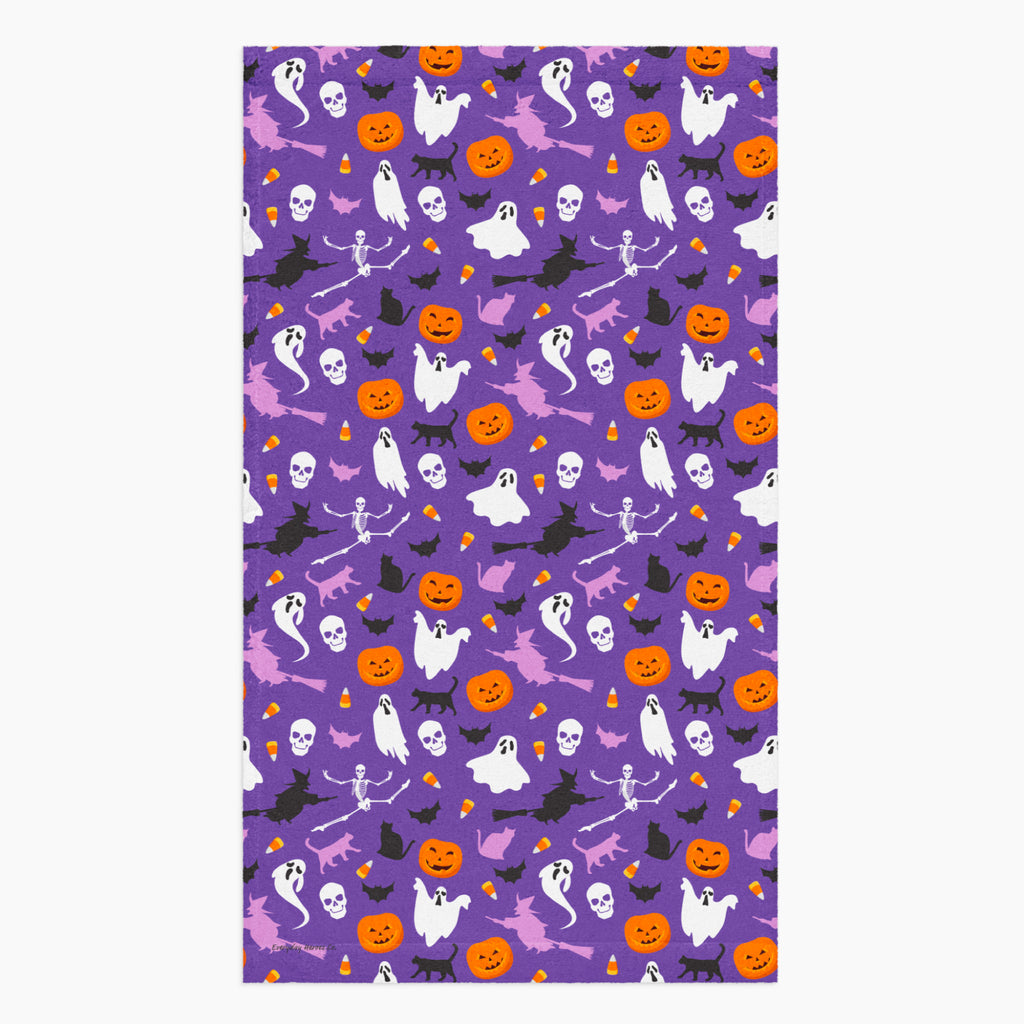 A rectangular cloth kitchen towel featuring a repeating Halloween pattern on a purple background, with white ghosts, black cats, orange pumpkins, and small candy corns.