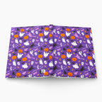 A hardcover notebook or journal shown open flat against a light gray background. The cover features a repeating Halloween pattern with purple background, orange pumpkins, white ghosts, black bats, and small yellow stars.