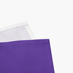 A close-up of a purple cloth napkin showing its solid purple front and a white reverse side with a stitched edge, set against a plain light-grey background.