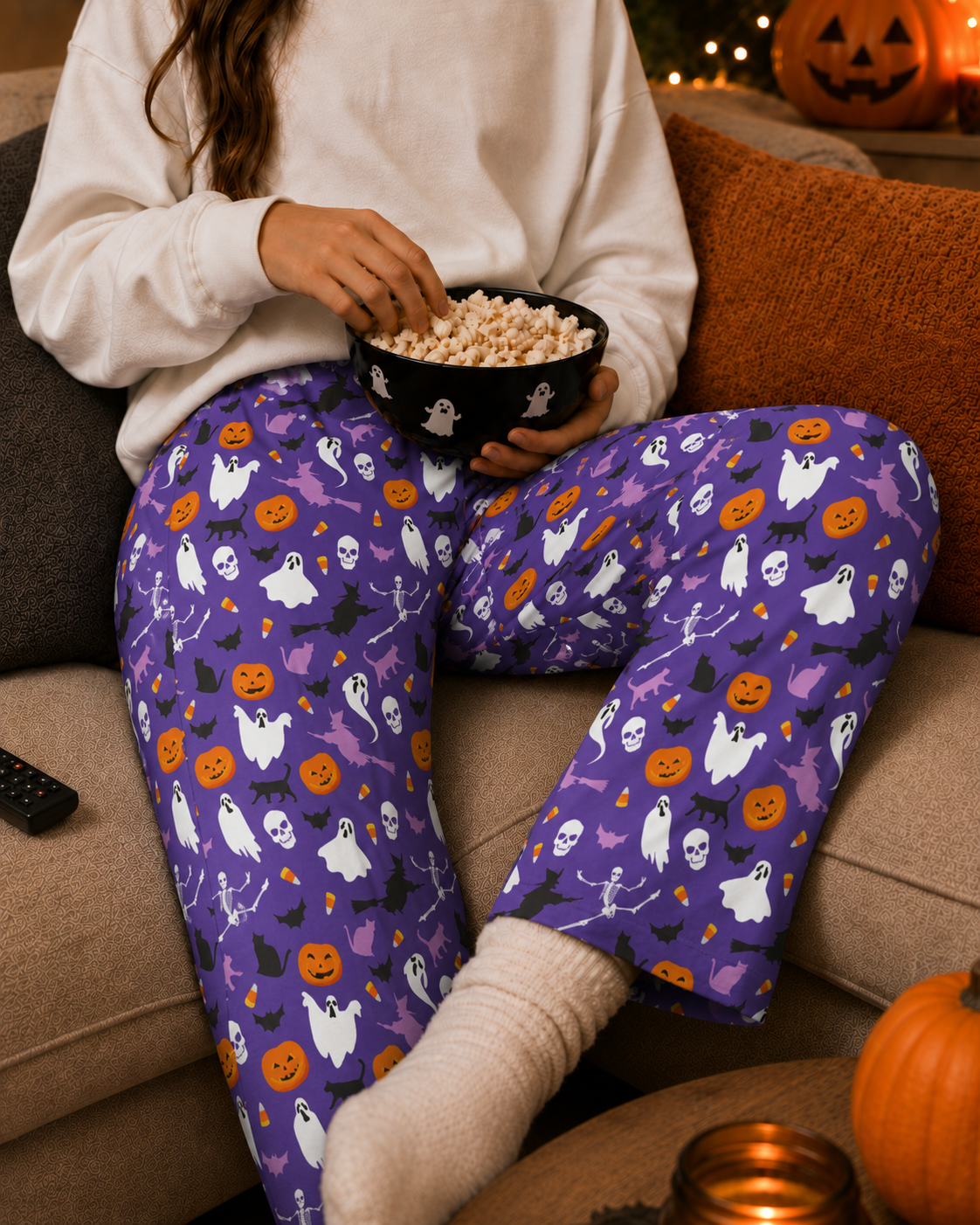 A person sitting on a couch wearing purple Halloween pajama pants with a ghost, pumpkin, bat, and skeleton print, holding a bowl of popcorn in a cozy living room setting.