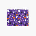 A rectangular cloth placemat featuring a repeating Halloween pattern on a purple background, with white ghosts, black cats and skeletons, orange pumpkins, and witches, shown on a light gray background.