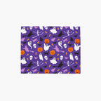 A rectangular cloth placemat featuring a repeating Halloween pattern on a purple background, with white ghosts, black cats and skeletons, orange pumpkins, and witches, shown on a light gray background.