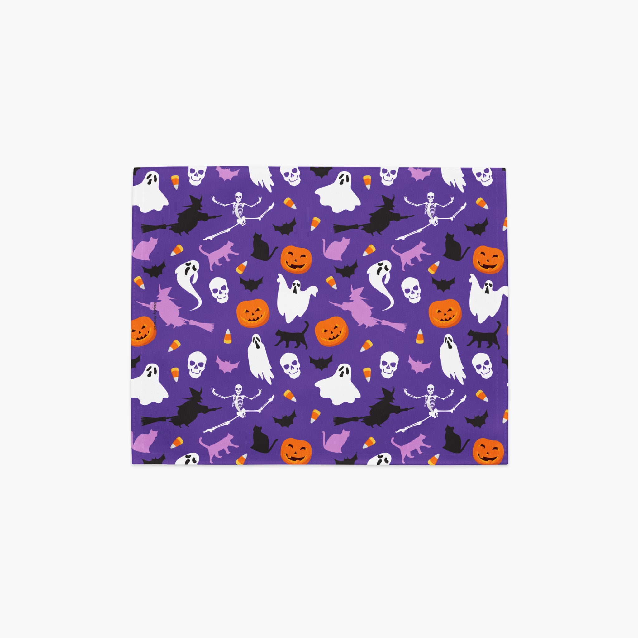 A rectangular cloth placemat featuring a repeating Halloween pattern on a purple background, with white ghosts, black cats and skeletons, orange pumpkins, and witches, shown on a light gray background.