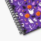 A spiral-bound notebook with a purple cover decorated with a Halloween pattern of pumpkins, ghosts, skeletons, skulls, bats, and candy corn, shown on a light-grey background.