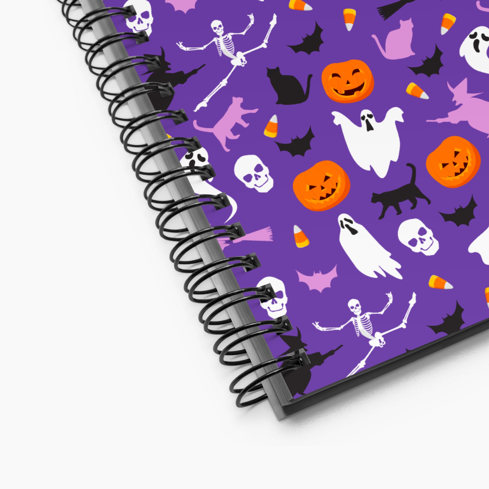 A spiral-bound notebook with a purple cover decorated with a Halloween pattern of pumpkins, ghosts, skeletons, skulls, bats, and candy corn, shown on a light-grey background.