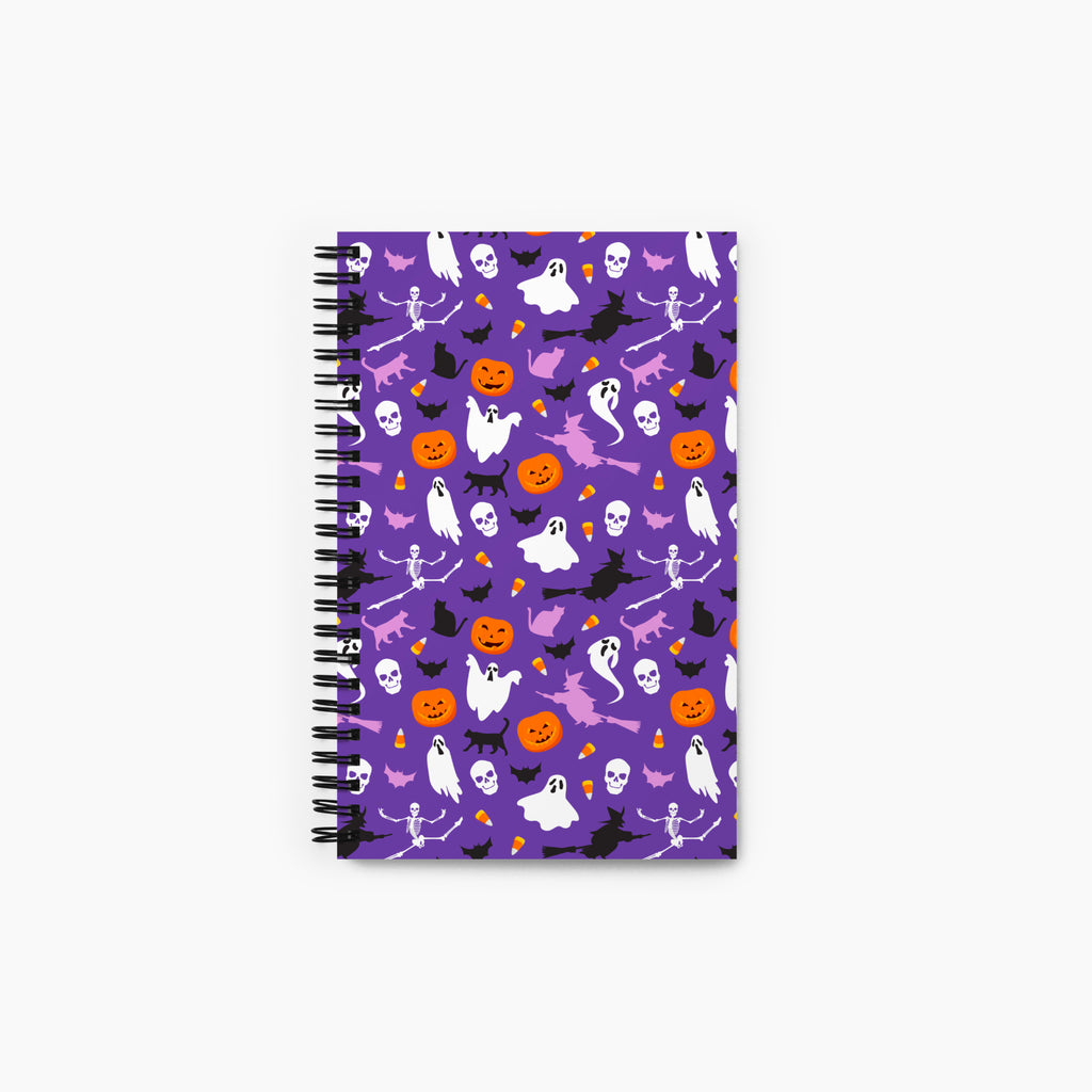 A spiral-bound notebook with a purple cover decorated with a repeating Halloween pattern of ghosts, pumpkins, bats, skulls, and witches, shown on a light gray background.