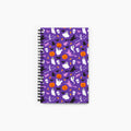 A spiral-bound notebook with a purple cover decorated with a repeating Halloween pattern of ghosts, pumpkins, bats, skulls, and witches, shown on a light gray background.