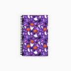 A spiral-bound notebook with a purple cover decorated with a repeating Halloween pattern of ghosts, pumpkins, bats, skulls, and witches, shown on a light gray background.