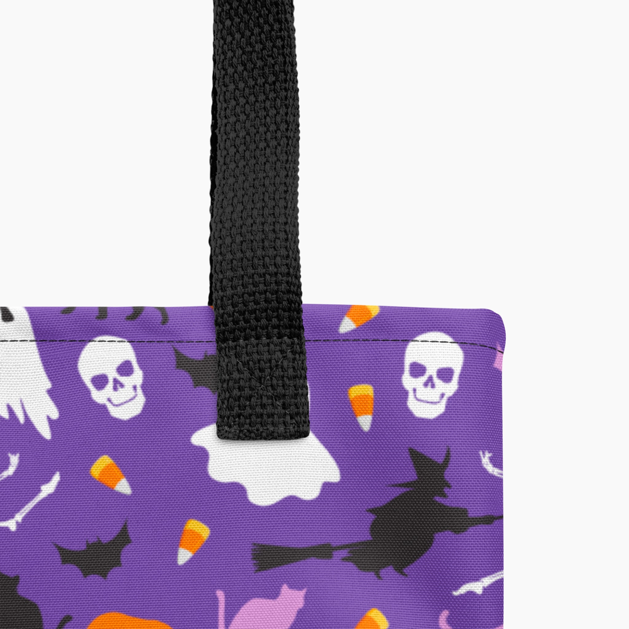 Close-up of a purple tote bag with a black woven handle and a Halloween print featuring white skulls, black bats, witch silhouettes, candy corn, a white ghost, and cat shapes on a light gray background.