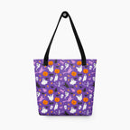 A purple tote bag with a Halloween pattern featuring white ghosts, orange pumpkins, black bats, witch silhouettes, and skulls, complete with black shoulder straps, shown against a plain light grey background.