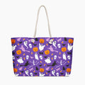 A purple tote bag with a Halloween-themed print featuring ghosts, pumpkins, bats, skeletons, and cats, complete with white rope handles, shown against a plain light-grey background.