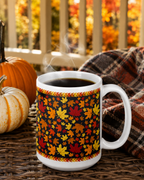 A white ceramic coffee mug with a black background and an all-over autumn leaf pattern in red, orange, yellow, and green, filled with steaming black coffee on a wicker table next to pumpkins and a plaid blanket.
