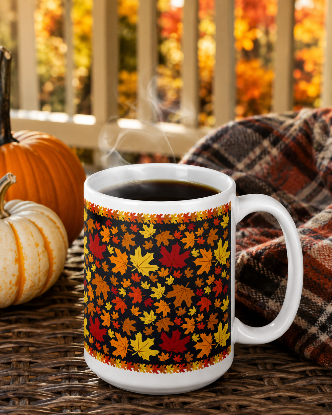 A white ceramic coffee mug with a black background and an all-over autumn leaf pattern in red, orange, yellow, and green, filled with steaming black coffee on a wicker table next to pumpkins and a plaid blanket.