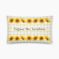 A rectangular throw pillow with a white and pale yellow gingham pattern, decorated with yellow sunflower illustrations and the phrase 'Follow the Sunshine' in green text, staged on a light grey studio background.