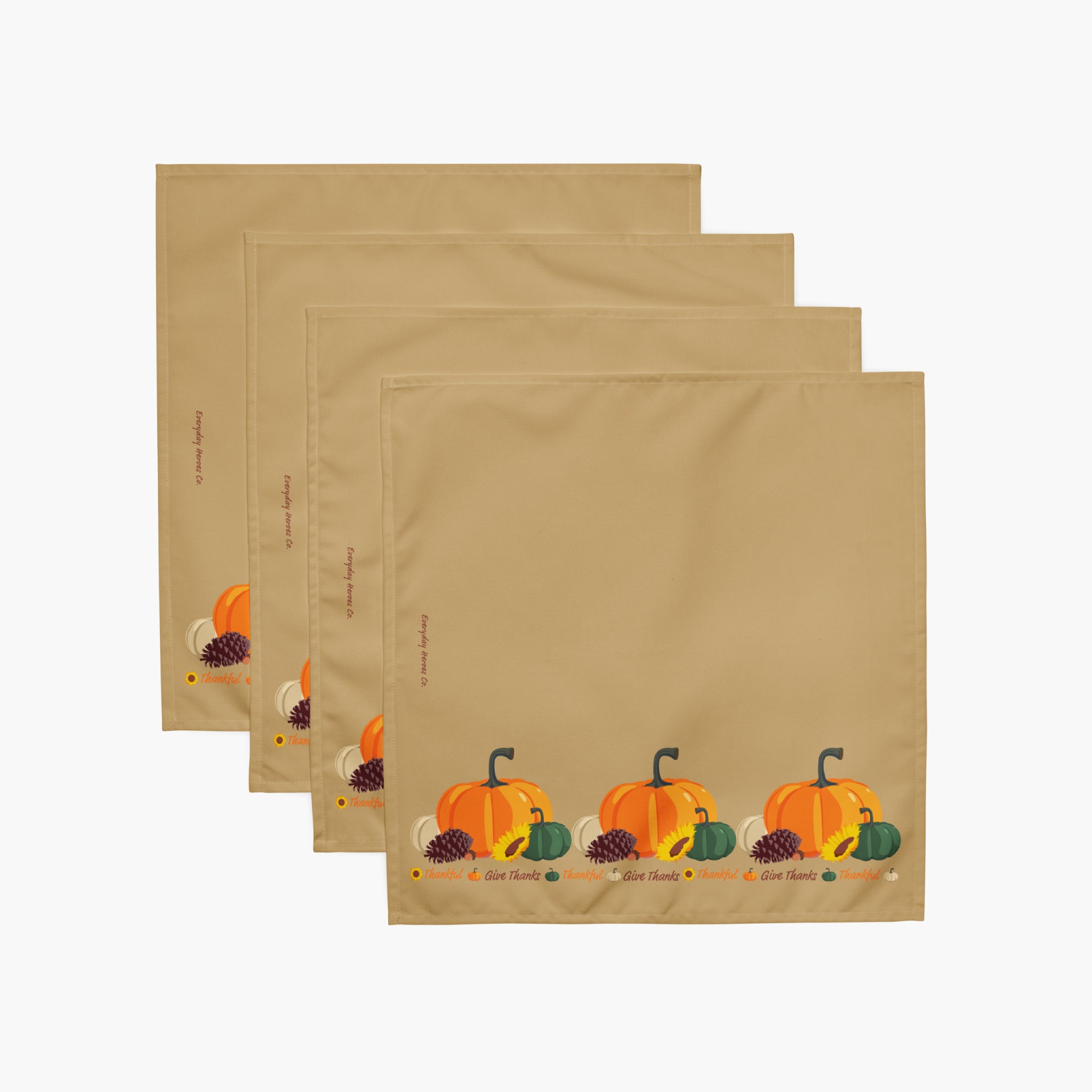 Four tan square cloth napkins layered in a fan arrangement, each printed with a small autumn harvest motif including orange pumpkins, green gourds, pinecones near the bottom.