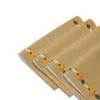 Close-up of four tan cloth placemats with a decorative autumn border featuring pumpkins and sunflowers, arranged in a staggered stack on a light grey background.