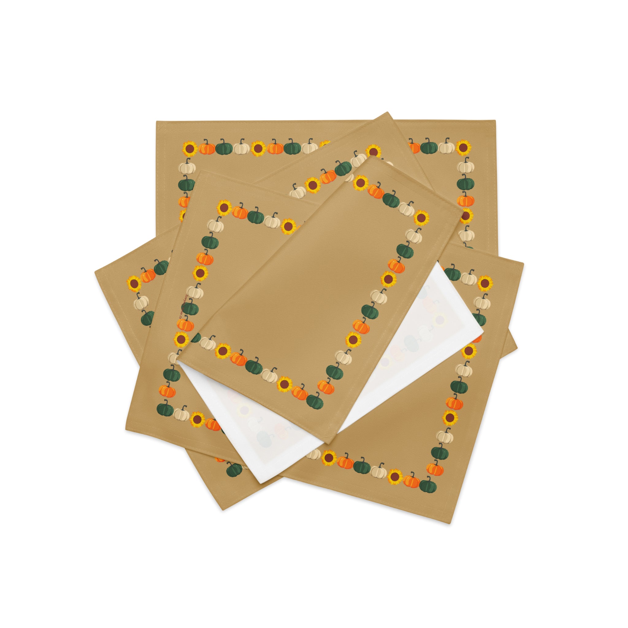 Close-up of four tan cloth placemats with a decorative autumn border featuring pumpkins and sunflowers, arranged in a stack on a light grey background.