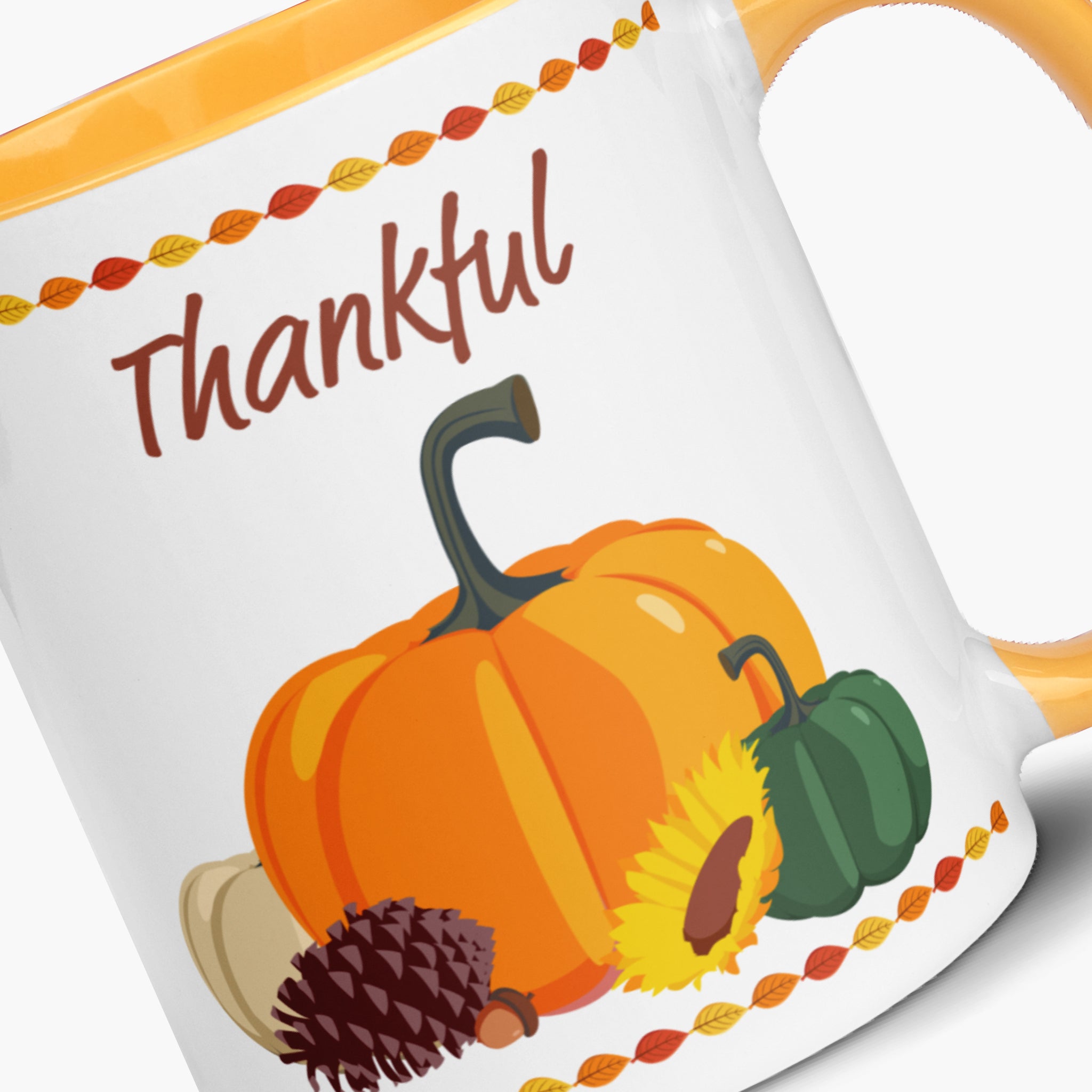 A close-up of a white ceramic mug with a yellow handle and interior, printed with the word 'Thankful' in brown script above an autumn harvest illustration featuring an orange pumpkin, green pumpkin, sunflower, pinecone, and decorative red/orange/yellow leaf borders.