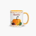 A white ceramic mug with a yellow-orange handle and interior, decorated with a fall harvest theme featuring pumpkins, a pinecone, a sunflower, and the word 'Thankful' in brown script.