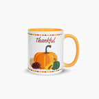A white ceramic mug with a yellow-orange handle and interior, decorated with a fall harvest theme featuring pumpkins, a pinecone, a sunflower, and the word 'Thankful' in brown script.