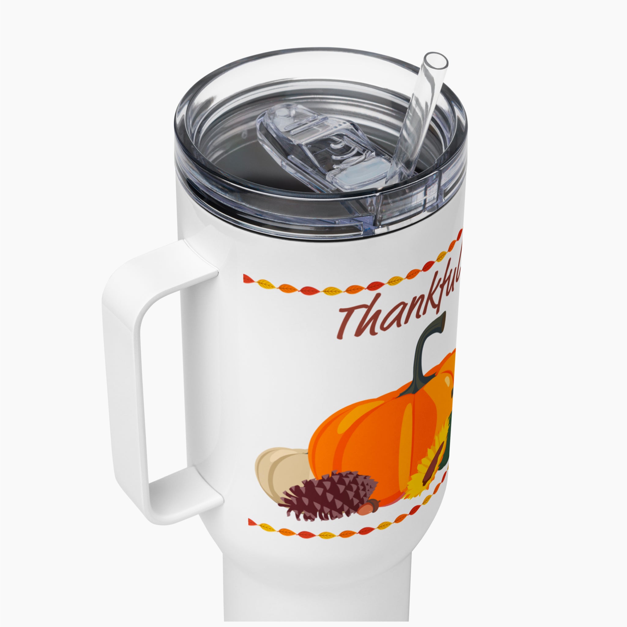 A close-up of a white insulated travel tumbler with a clear lid and straw, decorated with a Thanksgiving theme featuring an orange pumpkin, a pinecone, acorns, and the word 'Thankful' in brown script.