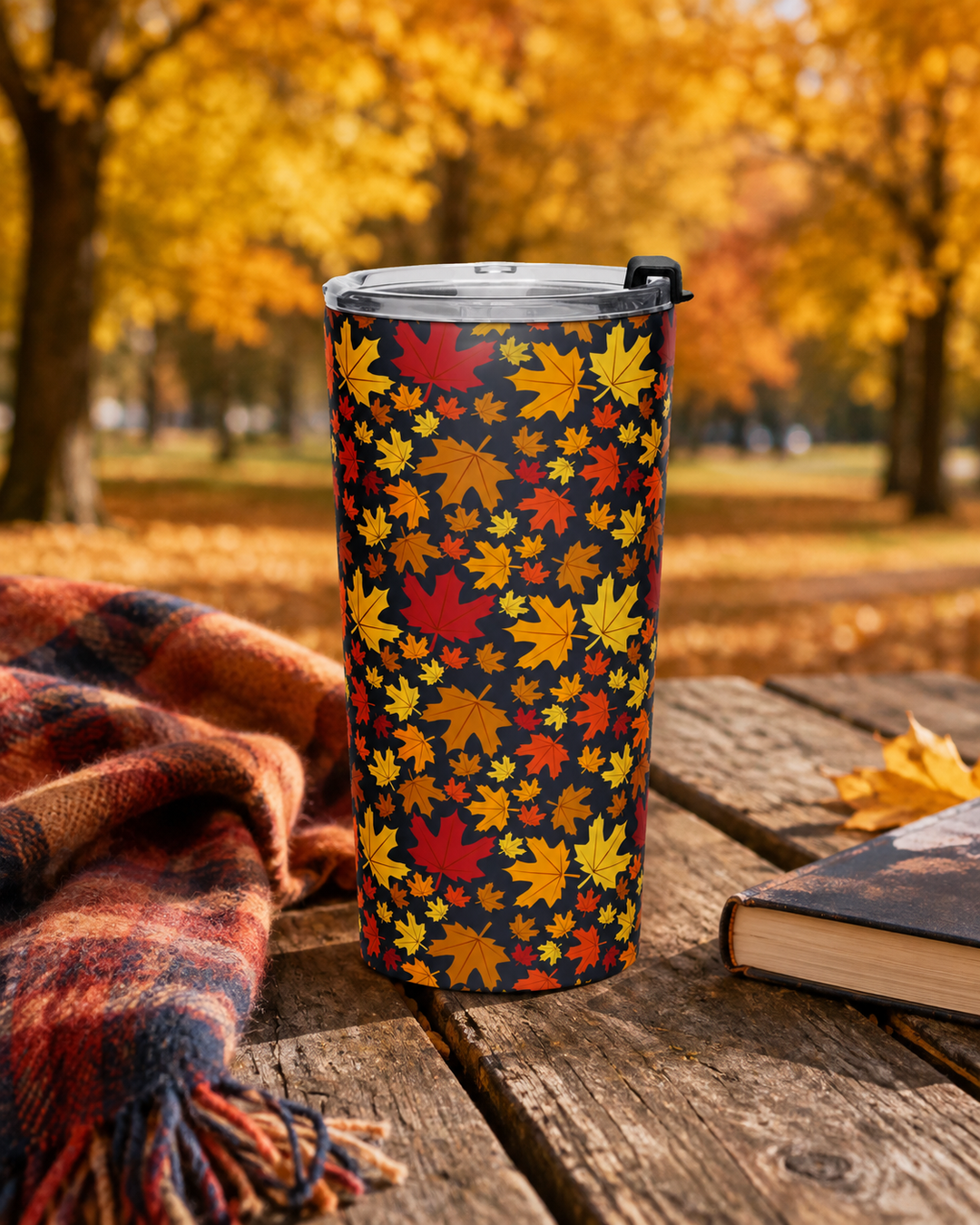 A tall insulated tumbler with a lid stands on a rustic wooden table outdoors. The tumbler has an all-over autumn leaf print in red, orange, yellow, and brown on a dark background.