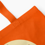 A closeup of an orange canvas tote bag with orange handles, featuring a 'Witch way to the Candy' message and cartoon girl dressed up as a witch on a broom, set against a plain light grey background.